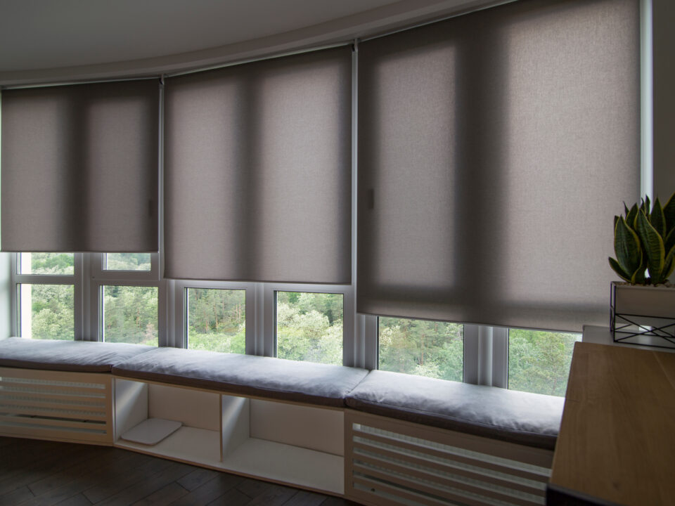 Motorized roller shades in the interior. Automatic roller blinds beige color on big glass windows. Home luxury curtains are above the windowsill with pillows. Summer. Green trees outside.