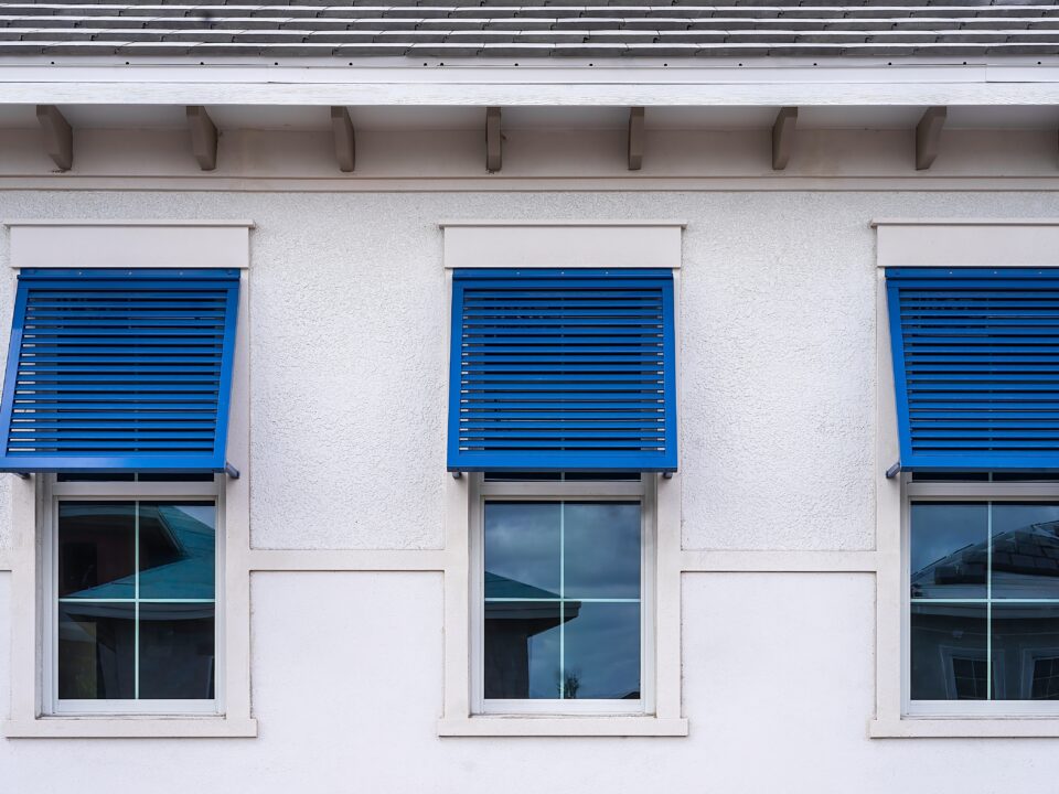 Closeup of three identical blue bahama shutters on top halves of windows.