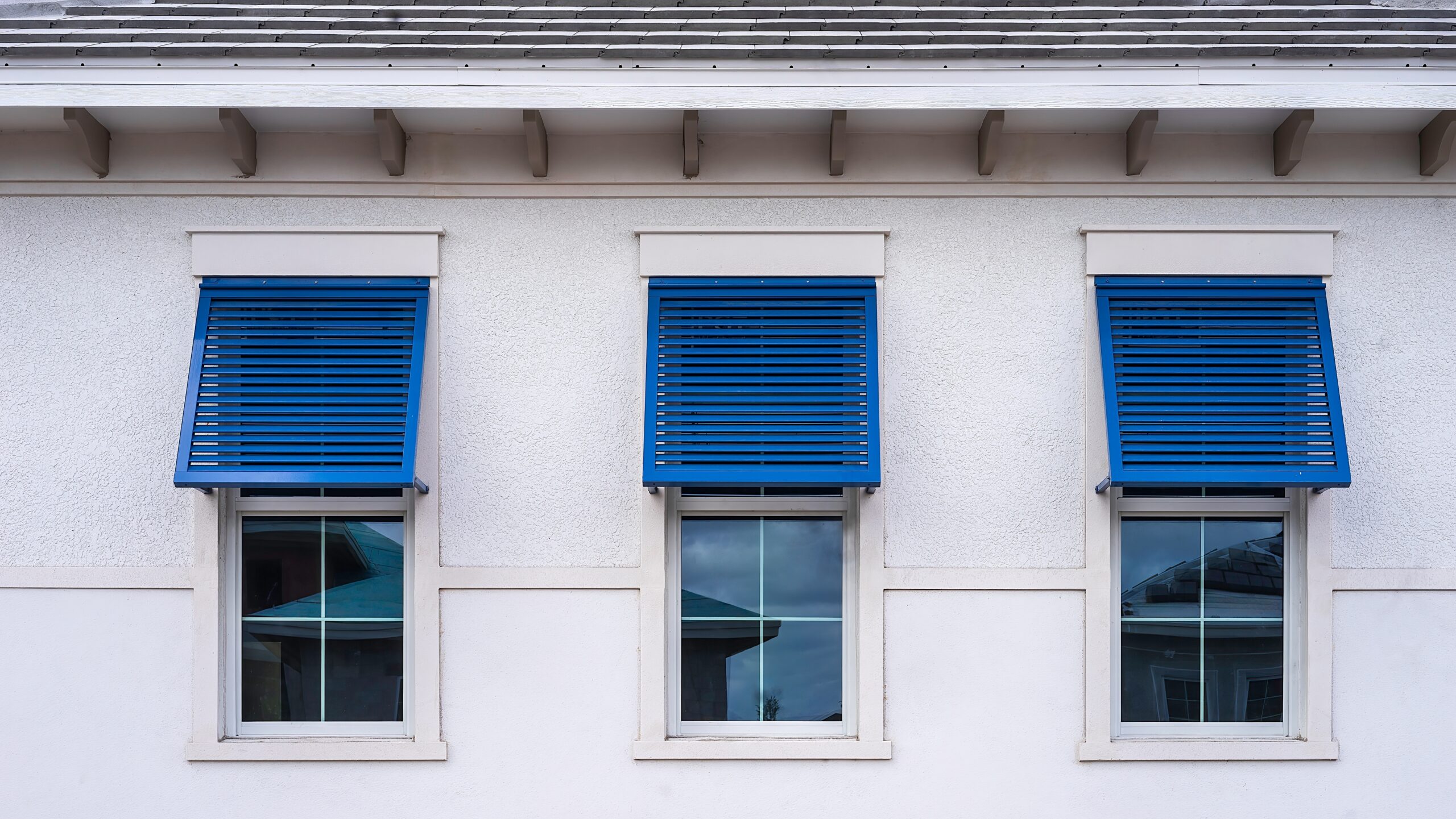Closeup of three identical blue bahama shutters on top halves of windows.